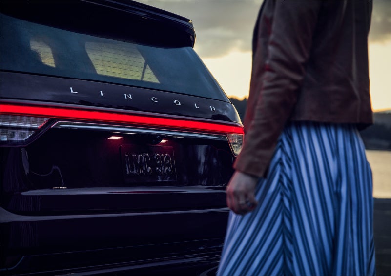A person is shown near the rear of a 2023 Lincoln Aviator® SUV as the Lincoln Embrace illuminates the rear lights | Pinnacle Lincoln in Nicholasville KY