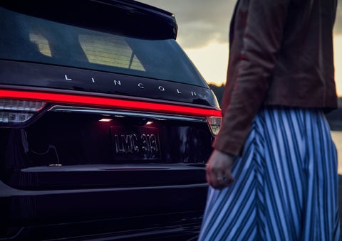 A person is shown near the rear of a 2024 Lincoln Aviator® SUV as the Lincoln Embrace illuminates the rear lights | Pinnacle Lincoln in Nicholasville KY